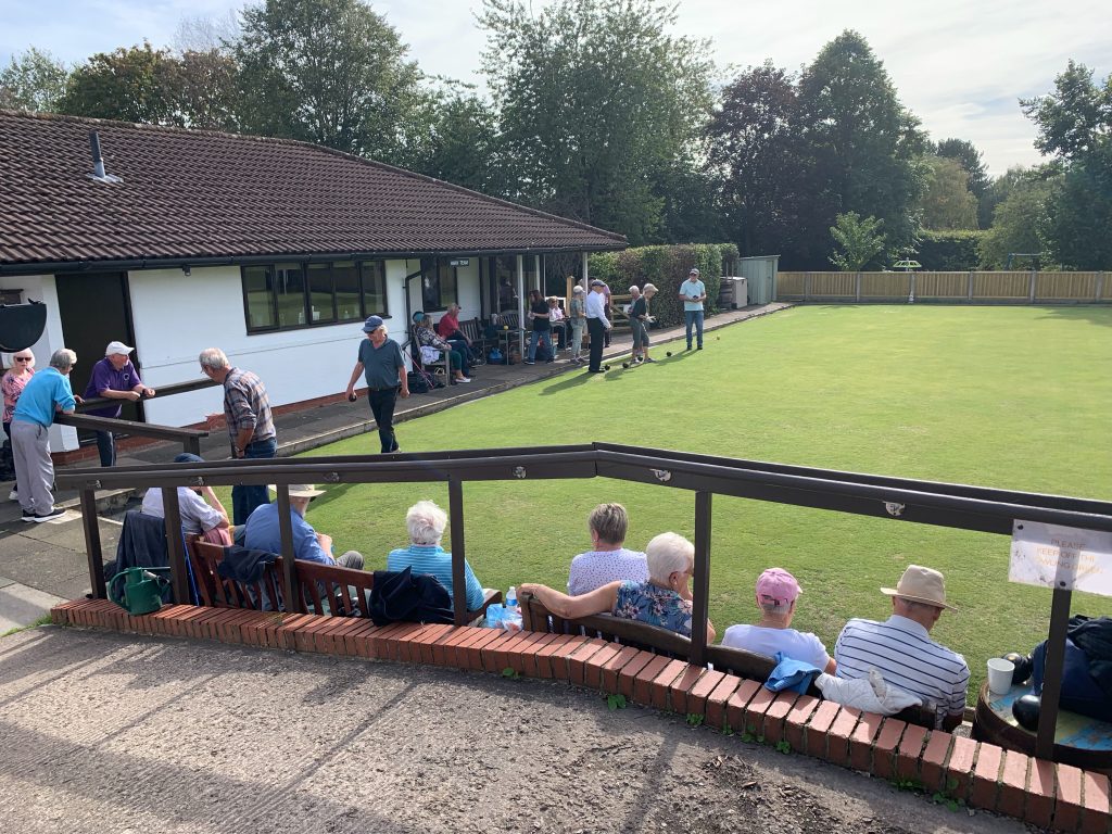 Bowlers on the green and spectators on benches. Taken from the ramp.